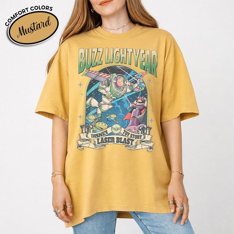 May include: Mustard-colored t-shirt featuring a graphic of Buzz Lightyear from Toy Story. The design includes the text "Buzz Lightyear", "Disney", "Laser Blast", and "Toy Story". The shirt has short sleeves and a crew neck.