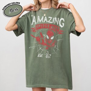 May include: Moss green t-shirt featuring a distressed graphic of Spider-Man with the text "AMAZING SPIDER-MAN" and "Est. '62". The shirt has a crew neck and short sleeves. The words "COMFORT COLORS" and "Moss" are in a circle in the upper left corner.