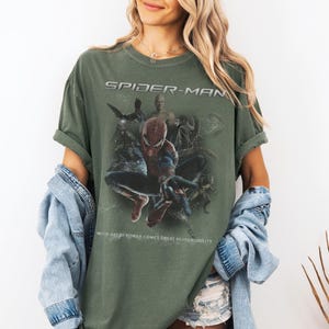 May include: An olive green t-shirt with a Spider-Man graphic. The text "SPIDER-MAN" is above the image, and the quote "WITH GREAT POWER COMES GREAT RESPONSIBILITY" is below. The graphic features Spider-Man and other characters.