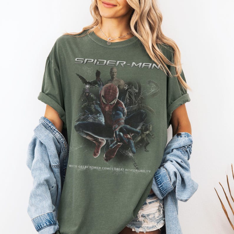 May include: An olive green t-shirt with a Spider-Man graphic. The text "SPIDER-MAN" is above the image, and the quote "WITH GREAT POWER COMES GREAT RESPONSIBILITY" is below. The graphic features Spider-Man and other characters.