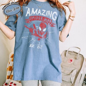 May include: Blue t-shirt featuring a distressed red and white graphic of Spider-Man with the text "THE AMAZING SPIDER-MAN" and "Est. '62". The shirt has short sleeves and a crew neck. A blue oval label in the upper left corner reads "COMFORT COLORS Blue Jean".