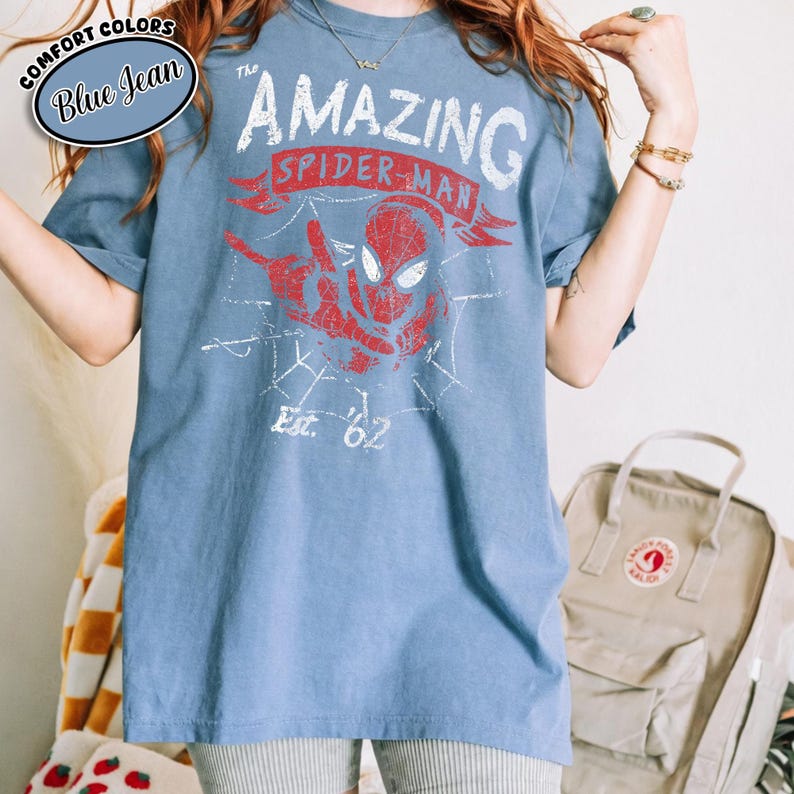May include: Blue t-shirt featuring a distressed red and white graphic of Spider-Man with the text "THE AMAZING SPIDER-MAN" and "Est. '62". The shirt has short sleeves and a crew neck. A blue oval label in the upper left corner reads "COMFORT COLORS Blue Jean".