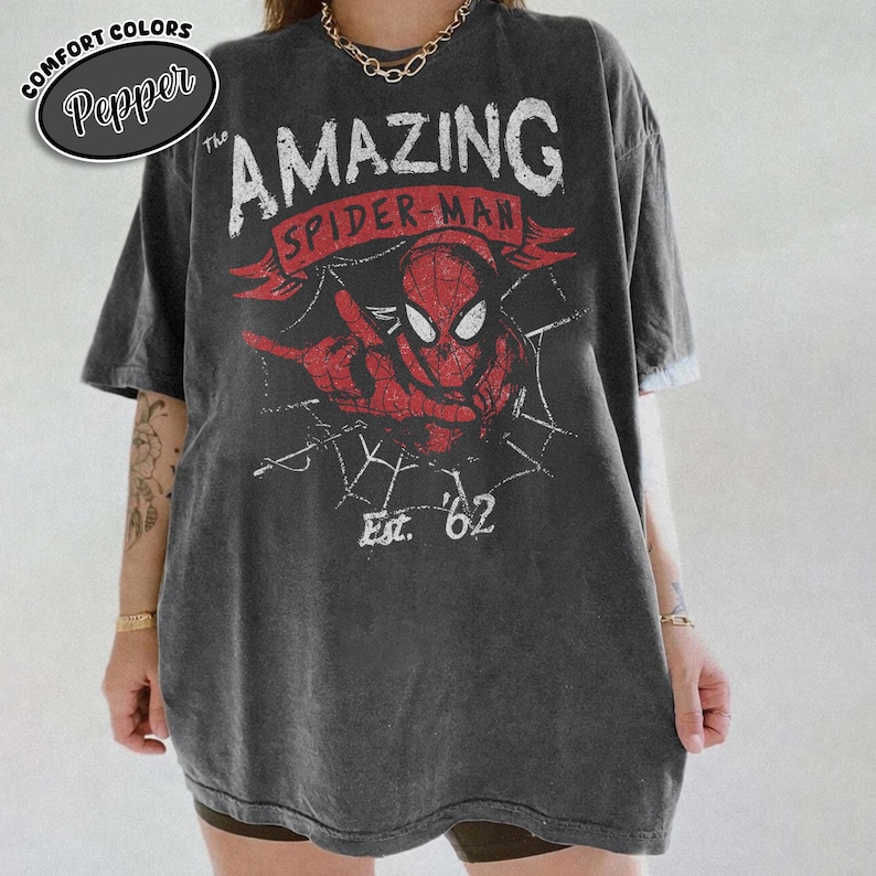 May include: Dark grey t-shirt featuring a distressed graphic of Spider-Man with the text "The Amazing Spider-Man" and "Est. '62" in red and white. The shirt has a vintage, worn look and a relaxed fit. The brand name "Pepper" is in a circle on the top left.