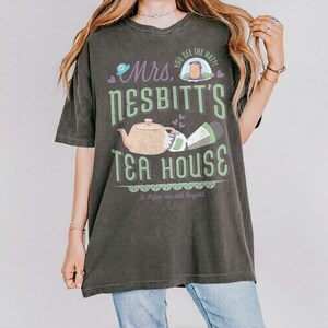 K&ouml;nnte beinhalten: Dunkelgraues T-Shirt mit einem gr&uuml;nen und lila Grafikdesign. Das Design zeigt den Text "Mrs. Nesbitt's Tea House", eine Teekanne, eine Tasse und den Satz "To Infini-tea and Beyond."