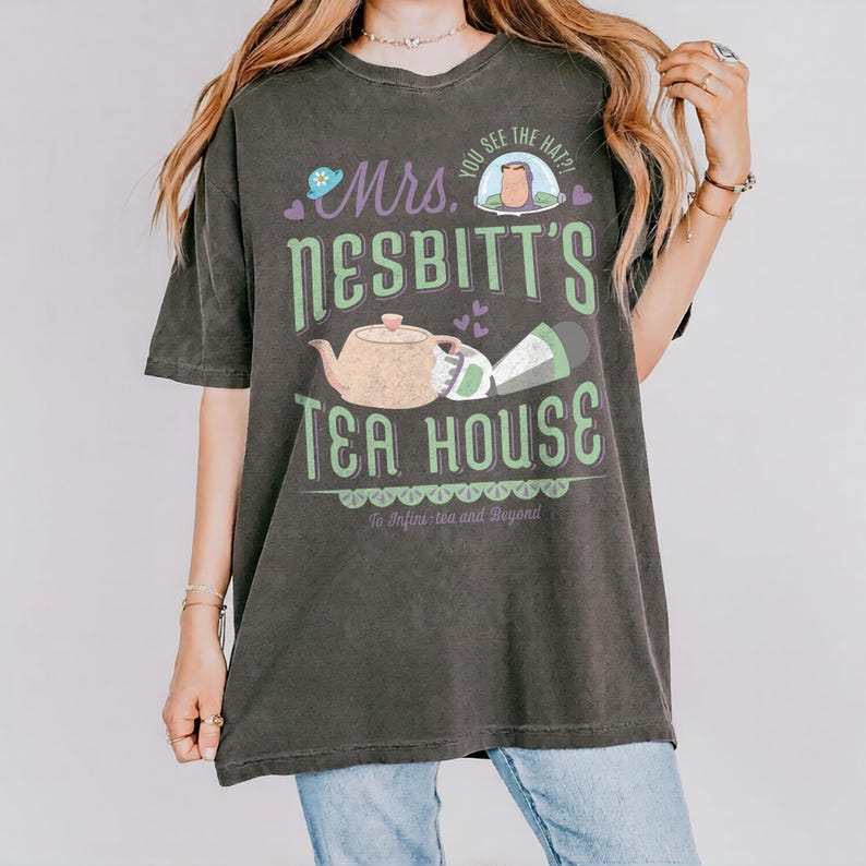 K&ouml;nnte beinhalten: Dunkelgraues T-Shirt mit einem gr&uuml;nen und lila Grafikdesign. Das Design zeigt den Text "Mrs. Nesbitt's Tea House", eine Teekanne, eine Tasse und den Satz "To Infini-tea and Beyond."