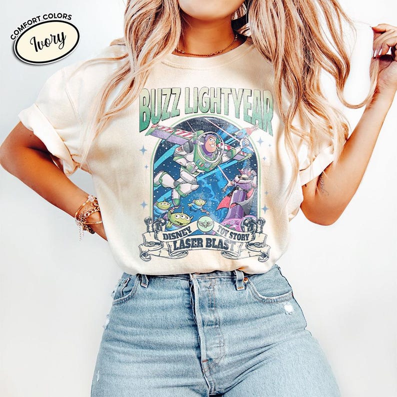 May include: Ivory-colored t-shirt featuring a graphic of Buzz Lightyear from Toy Story. The design includes the text "Buzz Lightyear", "Disney", "Toy Story", and "Laser Blast". The shirt has a crew neck and short sleeves.