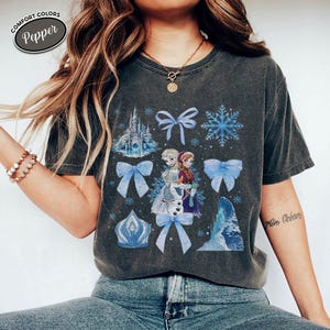 Puede incluir: Camiseta gris oscuro con un dise&ntilde;o de Elsa y Anna de Frozen, copos de nieve, lazos y un castillo. La camiseta tiene el texto "Comfort Colors Pepper" en un &oacute;valo blanco.