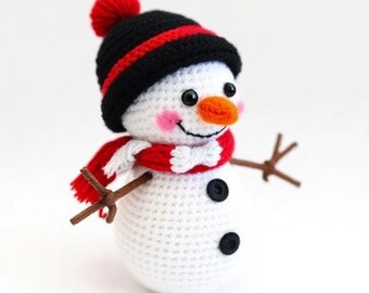 May include: A crocheted snowman with a black hat featuring a red stripe and a red pom-pom. The snowman has a red scarf, an orange carrot nose, black button eyes, and twig arms. Two black buttons are on the snowman's white body.