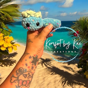 May include: A blue crocheted worm with a white crocheted crown and colorful sprinkles. The worm is being held in a hand with a tattoo of a skull and stars. The background is a beach scene with blue water and white sand. The text "Komfort by Kae Creations." is visible in the image.