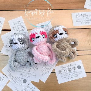 May include: Three crocheted sloth plushies in pink, gray, and brown. The plushies have white yarn for their eyes and mouths. The plushies are sitting on a wooden surface with business cards that say "Komfort by Kae Creations".