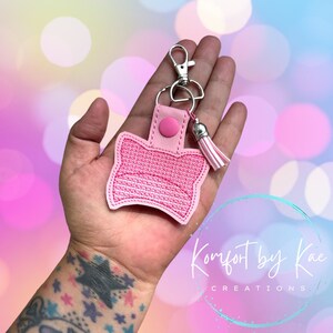 May include: Pink and white embroidered keychain with a tassel. The keychain is shaped like a cat's head and has a silver metal ring and clasp. The text "Komfort by Kae Creations" is visible in the background.