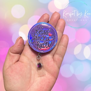May include: A round, purple badge reel with the phrase "Bless Your Heart" in cursive script. The badge reel has a holographic border and a clear plastic clip with a purple gem. The background is a soft gradient of pink, blue, and purple.