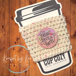 May include: A handmade, off-white crochet cup cozy with pink flecks, featuring a circular patch that reads "Bless Your Heart" in cursive. The cozy is wrapped around a coffee cup sleeve, with the words "Cup Cozy" and "100% Handmade" printed on it.