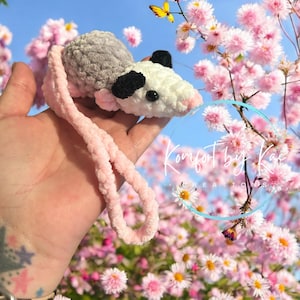 May include: A crocheted opossum plushie with a pink and gray color scheme. The plushie is holding a pink yarn loop and is in front of a background of pink flowers.