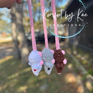 May include: Three handmade crochet cat toys shaped like mice, in white, gray, and brown, each with pink ears and a pink tail. The toys are suspended from a pink cord. The text "Komfort by Kae CREATIONS" is visible in the background.