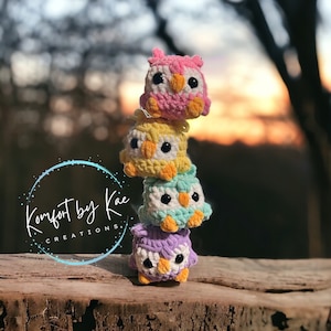 May include: A stack of four crocheted owl figurines in pink, yellow, teal, and purple. Each owl has white and orange accents, with black eyes. The owls are stacked on a wooden surface, with a blurred background of trees and sky.