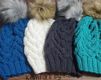 May include: Four cable-knit beanies in shades of teal, white, charcoal, and turquoise, each topped with a faux fur pom. The beanies are displayed on a wooden surface. The text "Komfort by Kae Creations" is visible.