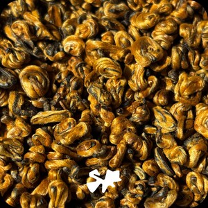 Könnte beinhalten: Eine Ansammlung von fest gerollten, getrockneten Teeblättern mit einer ausgeprägten Farbpalette aus tiefem Goldbraun und dunklem Anthrazit. Die Blätter sind zu kleinen, gleichmäßigen, spiralförmigen Clustern verarbeitet, die den Rahmen füllen.