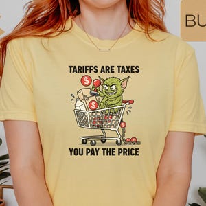 May include: Yellow t-shirt with the text "Tariffs are Taxes, You Pay the Price." The graphic shows a green goblin in a shopping cart filled with boxes and dollar signs. The shirt has short sleeves.