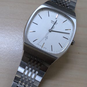 Puede incluir: Un reloj Seiko Quartz plateado con una esfera rectangular y una correa plateada. La esfera es blanca con líneas negras que marcan las horas y el logotipo de Seiko Quartz. La correa está hecha de pequeños eslabones rectangulares conectados.