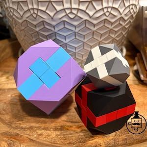 May include: Three geometric puzzle cubes in shades of purple, blue, gray, black, and red. The puzzles are stacked on a wooden surface, with a decorative white vase in the background. The cubes have interlocking designs.