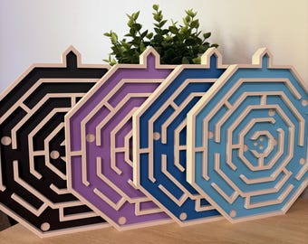 Puede incluir: Cuatro rompecabezas de laberinto de madera en negro, morado, azul y celeste. Cada rompecabezas tiene una combinación de colores única y un diseño de laberinto complejo. Los rompecabezas son octogonales con un borde claro.