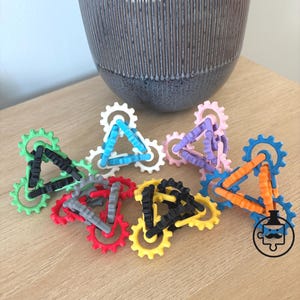 May include: A collection of colorful, interlocking gear-shaped toys. The gears are in various colors, including green, red, yellow, blue, and pink, and are connected by triangular frames. A decorative pot is in the background.