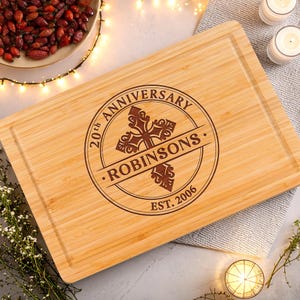 May include: A rectangular bamboo cutting board featuring a laser-engraved design. The center displays a decorative cross with the text ROBINSONS, surrounded by a circular border with the words 20th ANNIVERSARY and EST. 2006.