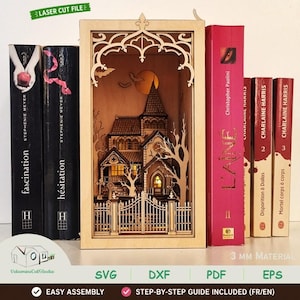 May include: A laser-cut wooden book nook insert featuring a multi-layered haunted house scene with a gated entrance, bare trees, bats, and a full moon. The structure is illuminated from within and positioned between several hardcover books on a shelf.