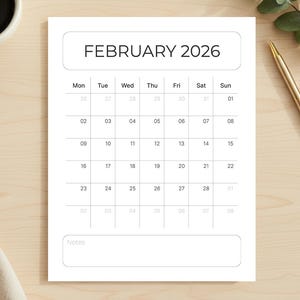 Puede incluir: Calendario blanco de febrero de 2026, sobre una superficie de madera clara. Hay una taza de café, un bolígrafo y vegetación. El calendario tiene un diseño sencillo.