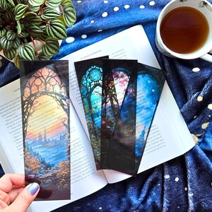 May include: Four decorative bookmarks with landscape scenes inside arched window frames. The bookmarks are on an open book, with a cup of tea. The bookmarks feature blue, orange, and purple hues. The scene includes trees, buildings, and a sky.