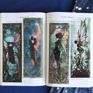 May include: An open book displaying four fairy-themed bookmarks. The bookmarks showcase silhouettes of fairies with detailed wings, set against a backdrop of swirling colours and floral patterns. The book rests on a blue surface.