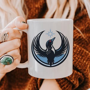 May include: White ceramic mug featuring a stylized black bird with outstretched wings, a red eye, and a blue star above. The bird is set within a blue circle. The mug has a white handle.