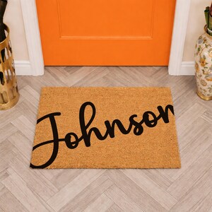 May include: A rectangular natural tan coir doormat featuring the name Johnson printed in a large, black, cursive script font across the center. The mat has a coarse, fibrous texture and is positioned horizontally on a light-colored floor.