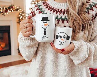 May include: Two white ceramic mugs with snowman faces. One mug has a snowman wearing a hat and tie, with the name "Robert" below. The other mug has a snowman with earmuffs and the name "Alice".