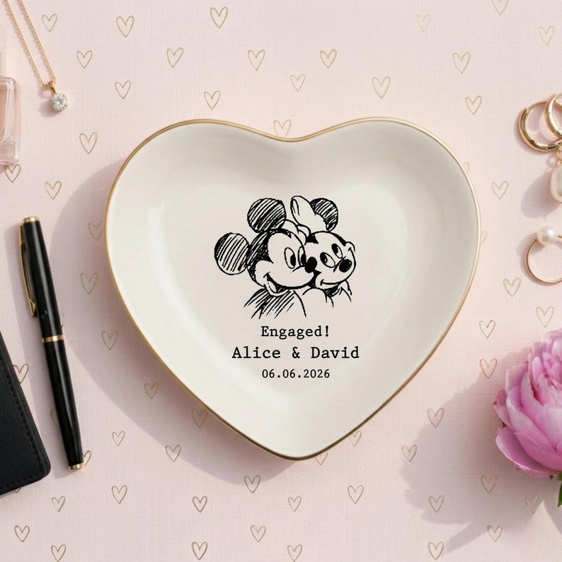 May include: A heart-shaped ceramic trinket dish with a gold-colored rim. The center features a black line-art illustration of two cartoon mice, with the text "Engaged! Alice & David" and the date "06.06.2026" printed in a black typewriter font.