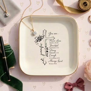 May include: A square, off-white trinket tray with a gold rim. The tray features a black cross design with floral accents and the name "Jennifer." Text includes "Never alone," "Beautiful," "Strong," "Enough," and "Always Loved."