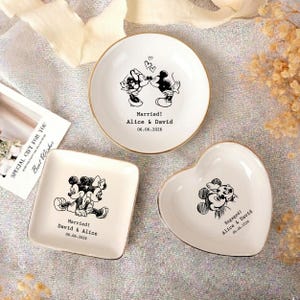 May include: Three white ceramic trinket dishes with gold rims. One round dish features Mickey and Minnie kissing with the text Married! Alice & David 06.06.2026. One square dish shows them sitting together with Married! David & Alice 06.06.2026. One heart dish shows them hugging with Engaged! Alice & David 06.06.2026.