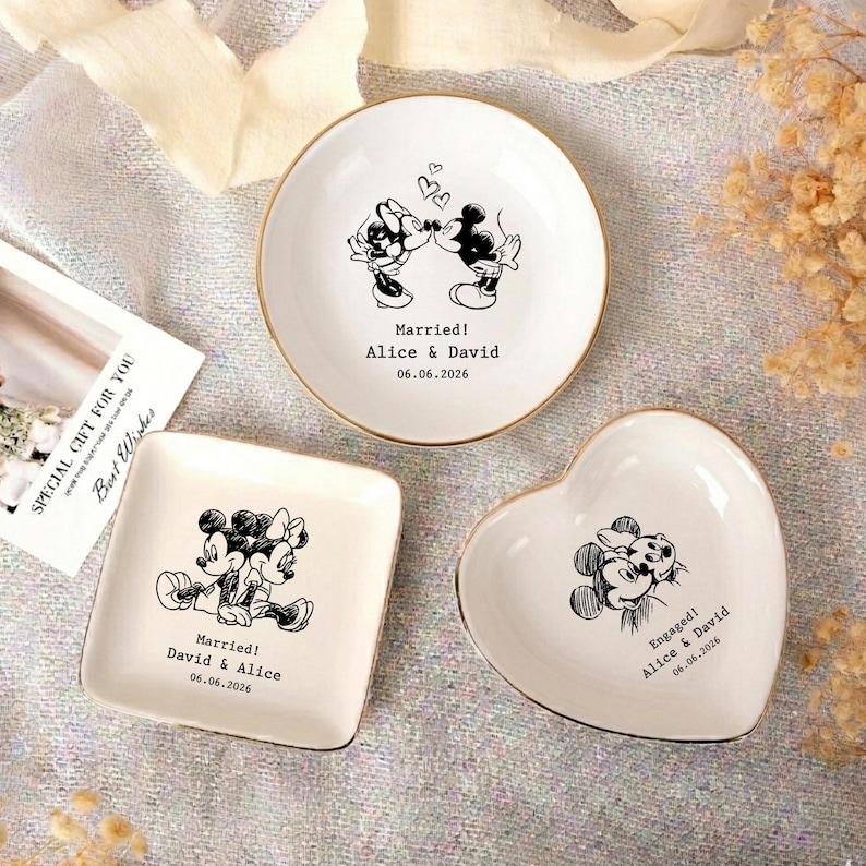May include: Three white ceramic trinket dishes with gold rims. One round dish features Mickey and Minnie kissing with the text Married! Alice & David 06.06.2026. One square dish shows them sitting together with Married! David & Alice 06.06.2026. One heart dish shows them hugging with Engaged! Alice & David 06.06.2026.