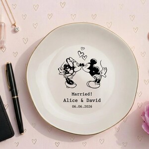 May include: A round white ceramic plate with a gold rim featuring a black line art illustration of two cartoon mice kissing under three small hearts. The text reads Married! Alice & David 06.06.2026 in a simple black typewriter font.