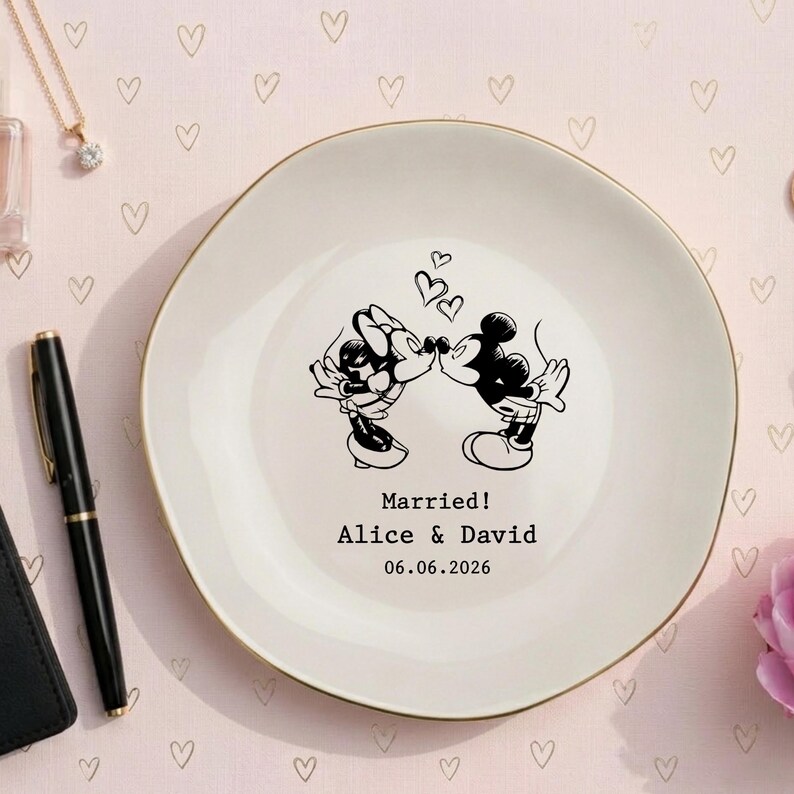 May include: A round white ceramic plate with a gold rim featuring a black line art illustration of two cartoon mice kissing under three small hearts. The text reads Married! Alice & David 06.06.2026 in a simple black typewriter font.