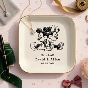 May include: A square off-white ceramic trinket dish with a gold-toned rim, featuring a black line-art illustration of Mickey and Minnie Mouse sitting back-to-back. The text reads Married! David & Alice 06.06.2026 in a black typewriter font.