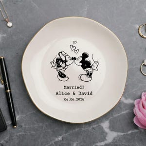 May include: A round white ceramic trinket dish with a gold-colored rim. The center features a black line art illustration of two cartoon mice kissing beneath three small hearts. Below the illustration is the text: Married! Alice & David 06.06.2026.