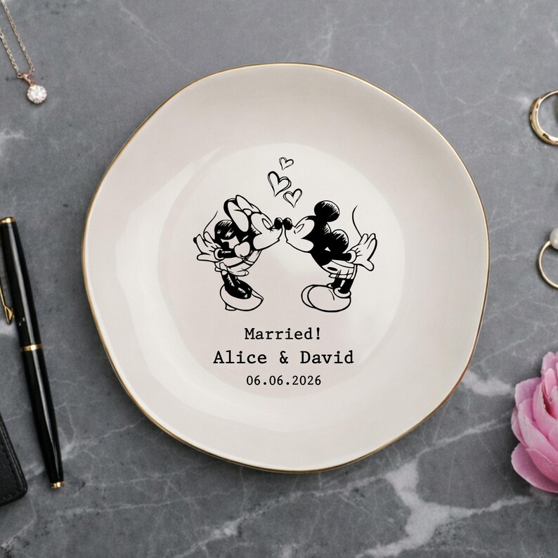 May include: A round white ceramic trinket dish with a gold-colored rim. The center features a black line art illustration of two cartoon mice kissing beneath three small hearts. Below the illustration is the text: Married! Alice & David 06.06.2026.