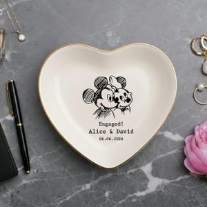May include: A heart-shaped ceramic trinket dish with a gold-colored rim. The center features a black line-art illustration of Mickey and Minnie Mouse, with the text "Engaged! Alice & David 06.06.2026" printed in a typewriter-style font.