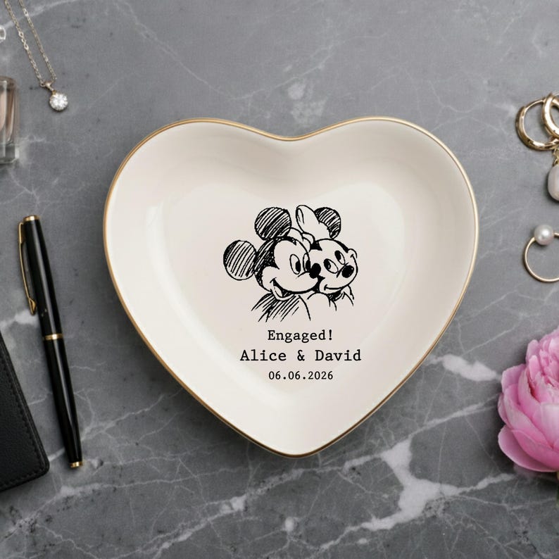 May include: A heart-shaped ceramic trinket dish with a gold-colored rim. The center features a black line-art illustration of Mickey and Minnie Mouse, with the text "Engaged! Alice & David 06.06.2026" printed in a typewriter-style font.