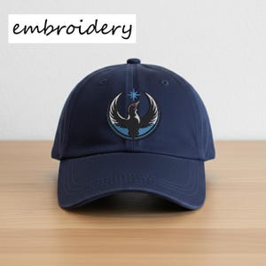 May include: Navy blue baseball cap with an embroidered emblem featuring a stylized bird with outstretched wings and a star. The word "embroidery" is printed in white on the top left. The cap is made of a durable fabric and has a curved brim.