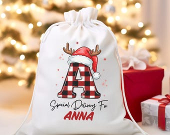 May include: A white drawstring gift bag with a festive design. The bag features a large letter "A" in a red and black buffalo plaid pattern, topped with a Santa hat and antlers. Text reads "Special Delivery For ANNA".
