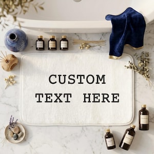 May include: A rectangular white bath mat with rounded corners featuring the black text CUSTOM TEXT HERE centered on the surface. The mat is surrounded by various bathroom accessories including small amber glass bottles and a navy blue towel.