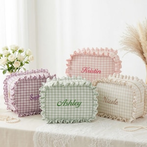 Puede incluir: Cuatro neceseres personalizados en colores pastel, cada uno con un estampado de cuadros y bordes con volantes. Los nombres bordados son: Kristin, Ashley, Nicole y Soph. Los neceseres se exhiben sobre una mesa blanca con detalles florales y decorativos.